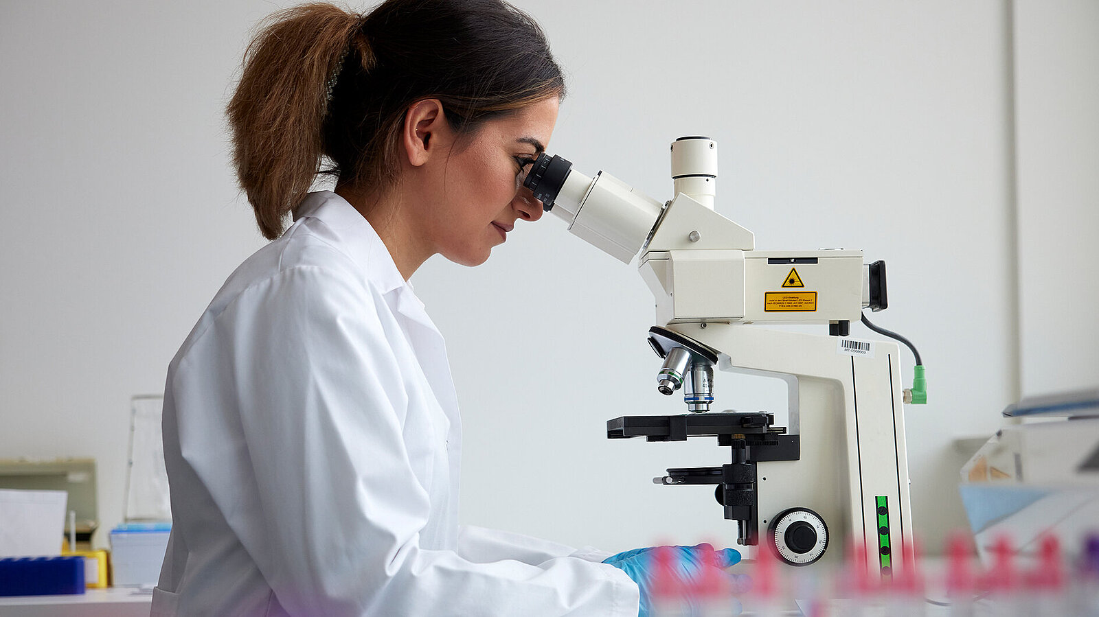 Eine junge Frau in Laborkittel und Handschuhen sieht durch ein Mikroskop.