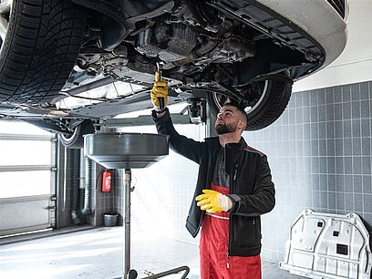 Ein junger Mann mit Handschuhen arbeitet am Unterboden eines Fahrzeugs.