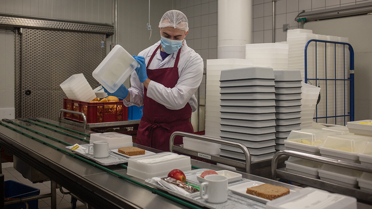 Ruben steht in Arbeitskleidung am Speiseverteilband und kontrolliert die Tabletts.