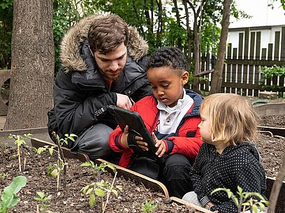 Ein junger Mann schaut mit zwei Kindern im Garten auf ein Tablet.