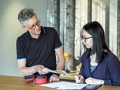 Ein Mann zeigt einer jungen Frau ein Blattt Papier während sie etwas in ihren Unterlagen notiert.