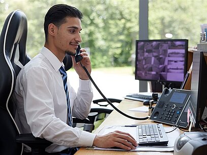 Ein junger Mann mit Krawatte telefoniert am Schreibtisch mit Überwachungsmonitor.