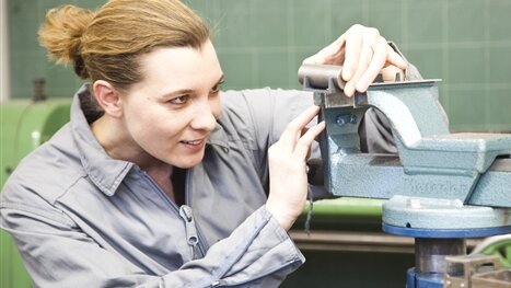 Eine junge Handwerkerin in Arbeitskleidung arbeitet in einem Werkraum an einer Maschine.
