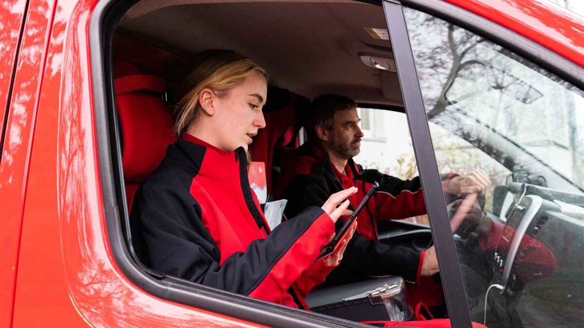 Eine Frau und ein Mann sitzen in Arbeitskleidung in einem Auto, die Frau tippt auf einem Tablet.