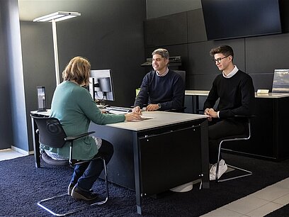 Zwei Männer beraten eine Frau an einem Schreibtisch.