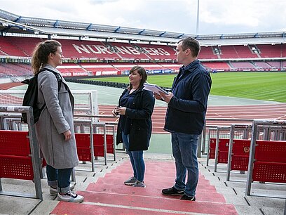 Eine junge Frau unterhält sich mit zwei Gästen auf der Tribüne eines Fußballstadions.