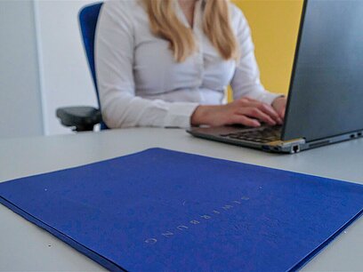 Eine Frau sitzt am Computer mit einer blauen Bewerbungsmappe im Vordergrund.