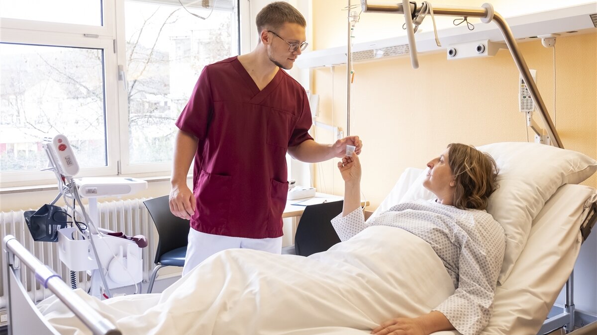 Ein junger Mann mit Pflegekleidung gibt einer Patientin im Krankenhausbett Medikamente.