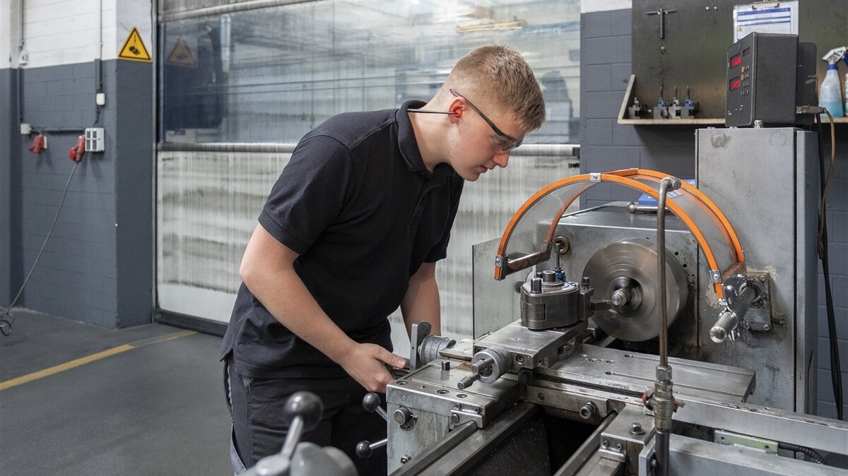 Ein junger Mann mit Schutzbrille bearbeitet ein Metallstück an einer Drehmaschine.