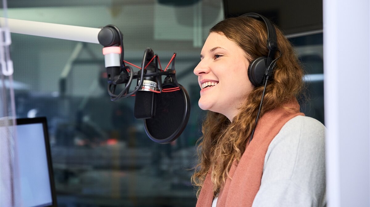 Eine junge Frau spricht in einem Tonstudio in ein Mikrofon und hat Kopfhörer auf.