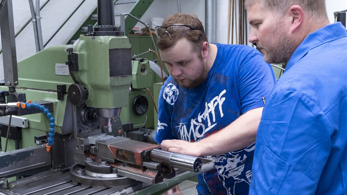 Ein Mann in Arbeitskleidung sieht einem jungen Mann bei der Arbeit an einer metallverarbeitenden Maschine zu.