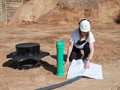 Eine junge Frau mit Schutzhelm überprüft in einer Baugrube Abwasserrohre.