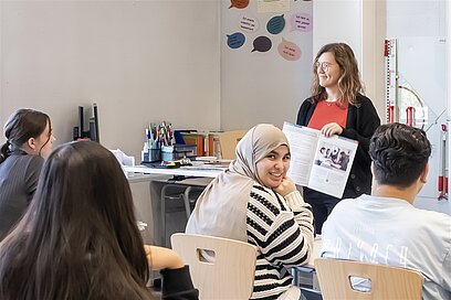 Eine Lehrerin zeigt Schülerinnen und Schülern im Klassenzimmer ein Heft.