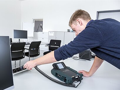 Ein Mann in Arbeitsschutzhose prüft die Steckdosen in einem Schulungsraum mithilfe eines Messgeräts.