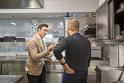 Ein Mann erklärt einem anderen Mann in der Küche etwas mit einem Plan in der Hand.