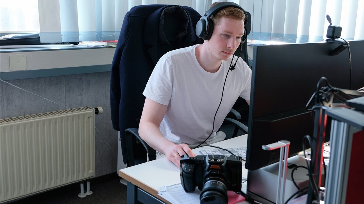 Ein junger Mediengestalter arbeitet mit Kopfhörer an seinem Computer, neben ihm liegt eine Kamera.