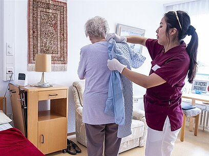Eine Pflegerin hilft einer älteren Dame beim Anziehen einer Jacke.