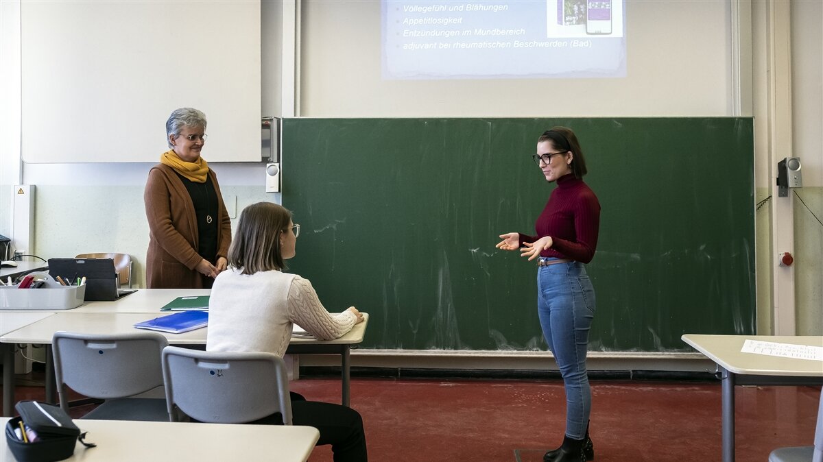 Eine junge Frau hält vor einer Frau und einer Schülerin einen Vortrag in einem Klassenzimmer.