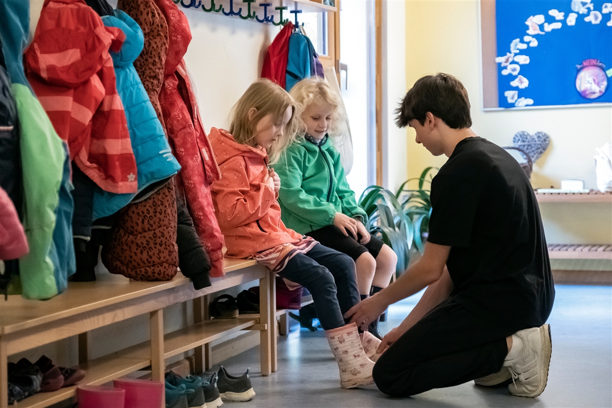 Ein junger Mann hilft zwei Kindern beim Schuhe anziehen.