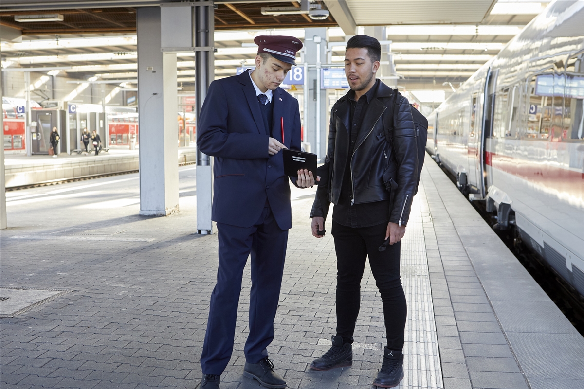 Ein Bahnmitarbeiter in Uniform und Mütze steht auf dem Bahnsteig eines Bahnhofs und spricht mit einem jungen Mann in schwarzer Kleidung. Der Mitarbeiter zeigt ihm etwas auf einem mobilen Gerät.