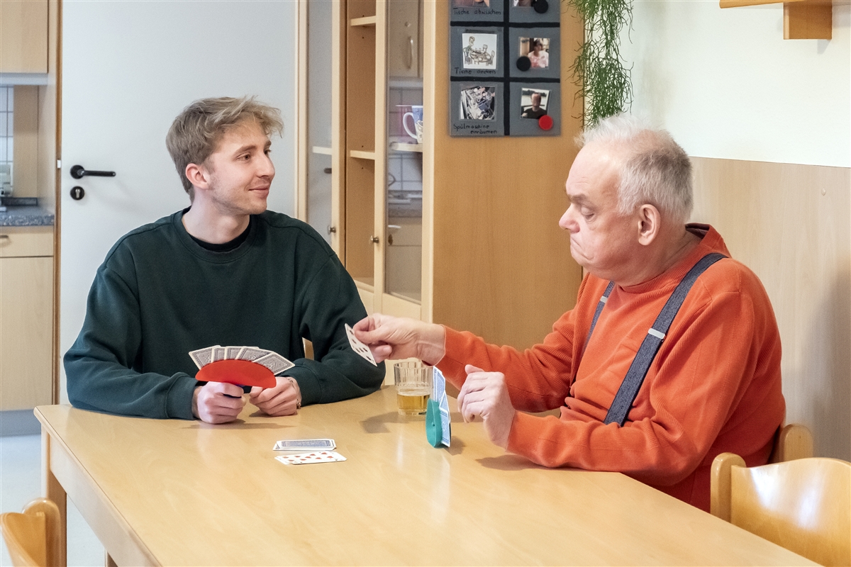Moritz spielt mit einem älteren Mann ein Kartenspiel am Tisch in einem Wohnraum.