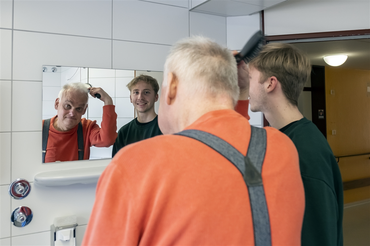 Moritz hilft einem älteren Mann, sich die Haare vor einem Spiegel zu kämmen.