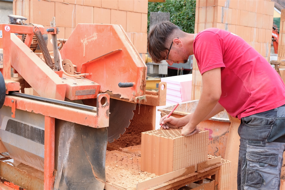 Adriatik steht auf der Baustelle an der Steinsäge und zeichnet mit einem Stift an, wo der Ziegelstein abgeschnitten werden soll.