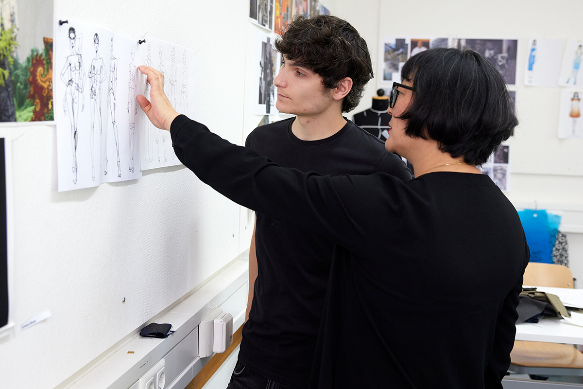 Pauls bespricht mit seiner Lehrerin seine Skizzen, die im Klassenzimmer an einer Wand hängen.