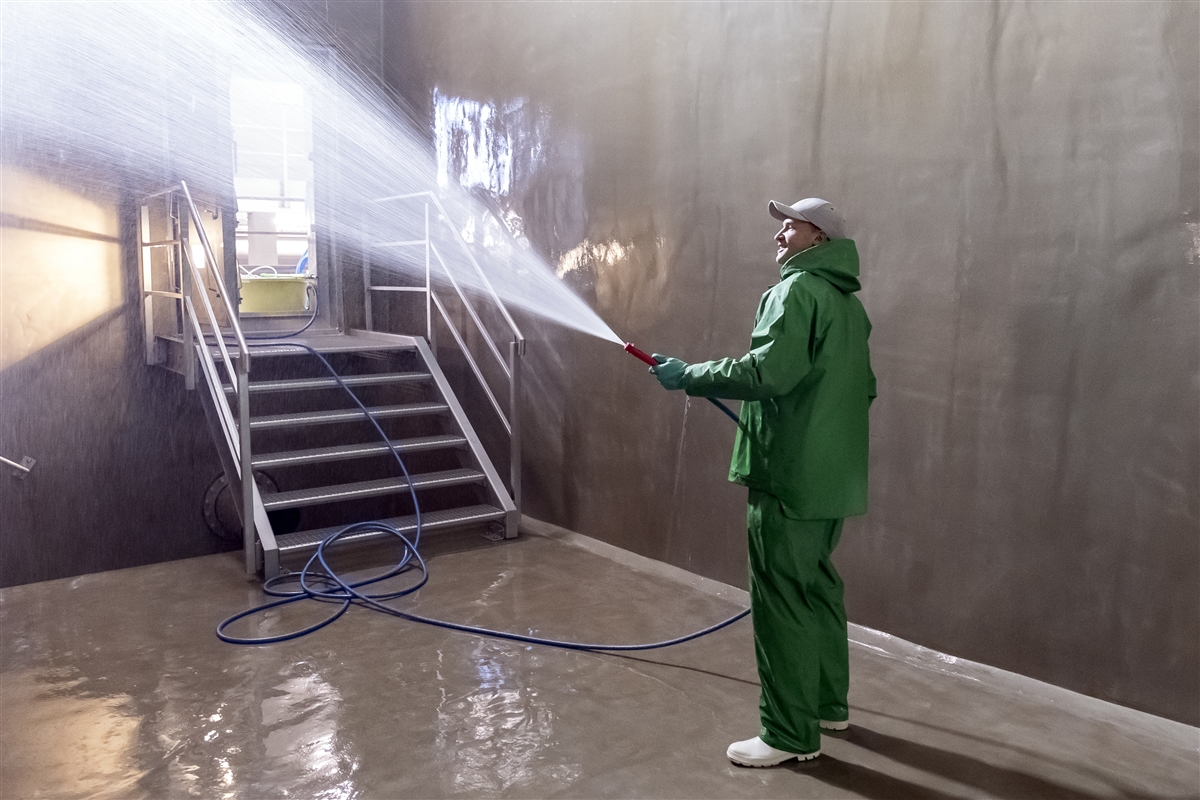 Daniel trägt einen wasserdichten Anzug und spritzt mit einem Schlauch Wasser in einen Raum aus Beton.
