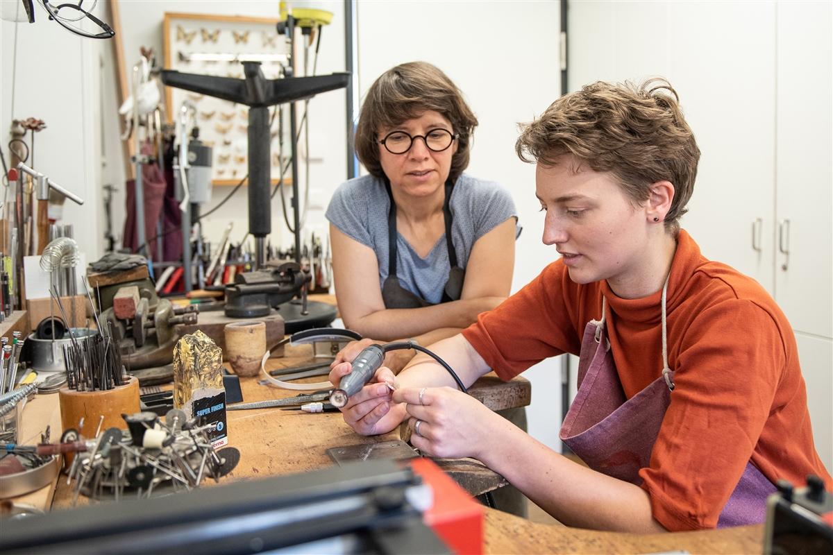 Eine Frau beaufsichtigt eine junge Frau beim Löten eines Metallstücks an einer Werkbank.