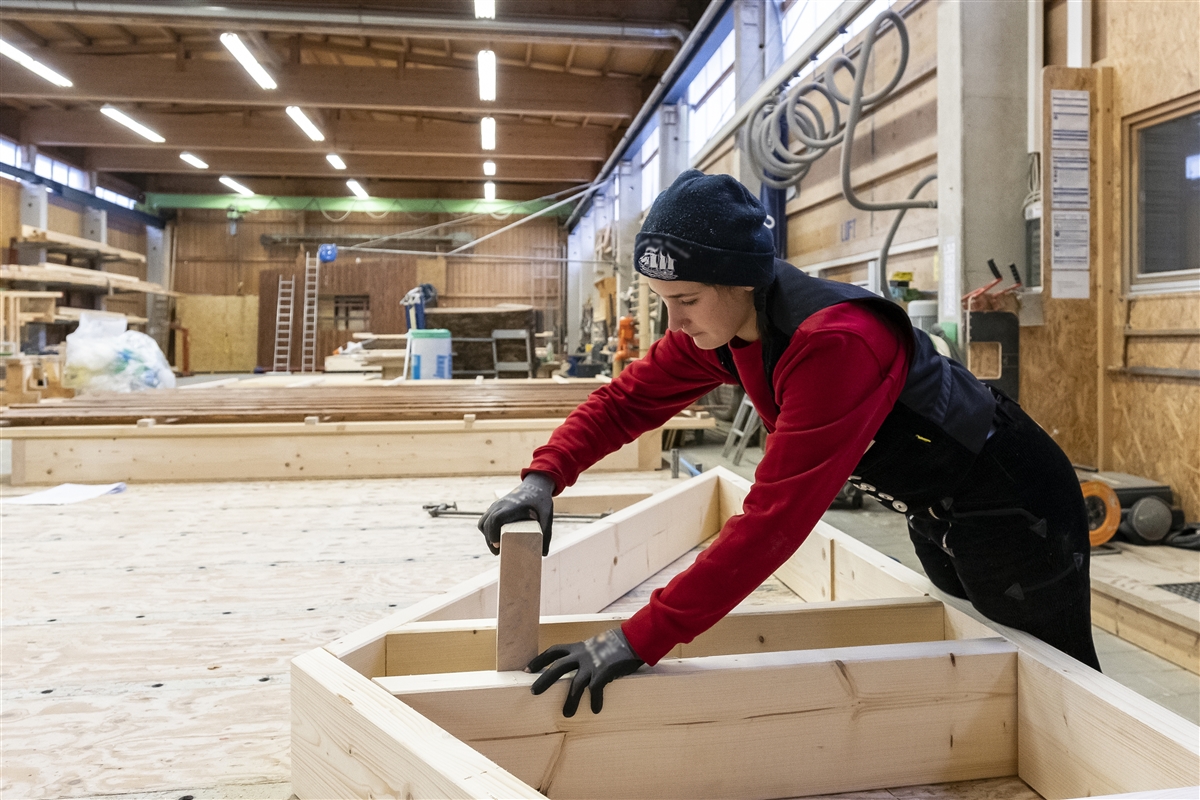 Eine junge Frau mit Schutzhandschuhen bearbeitet Holz in einer Werkstatt.